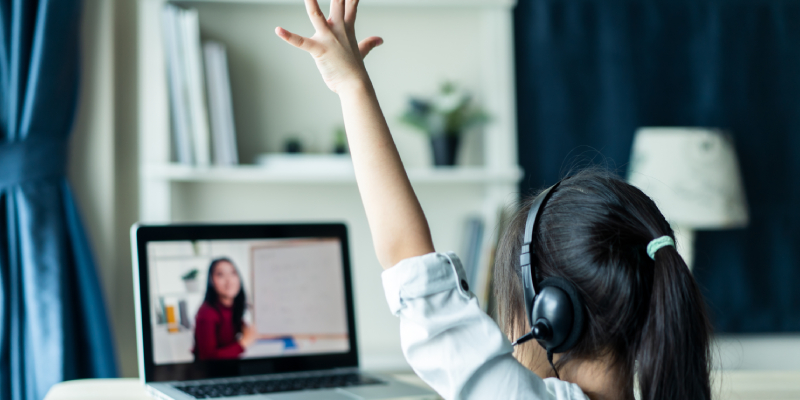 A teacher is taking online class and a girl is attending class from home. She is raising her hand, perhaps as an answer to a question.