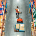 A worker carries boxes on a machine inside a warehouse filled with shelves stacked with boxes.