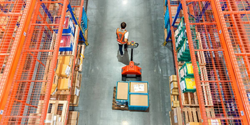 A worker carries boxes on a machine inside a warehouse filled with shelves stacked with boxes.