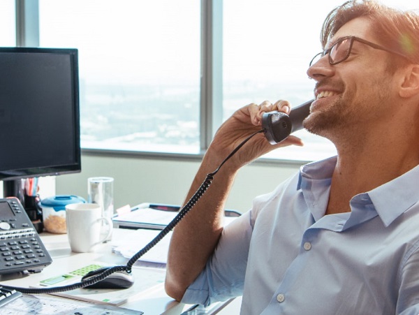 A man is talking on a landline phone from his office. A desktop is kept on the table.