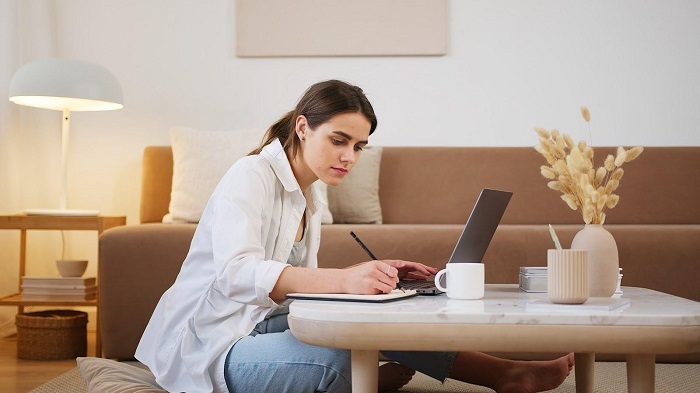 A girl is sitting in a drawing room writing something on a notepad on a table. In front of her a laptop is open.